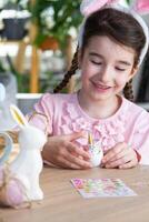 un' carino ragazza con rosa coniglietto orecchie fa un Pasqua mestiere - decora un uovo nel il modulo di un' unicorno con strass, corno, fiori nel il interno di un' Casa con impianti. foto