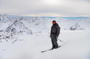 giovane uomo sta su un' roccia a il superiore di un' montagna con un' zaino e sci, Guardando un' lungo alpino panorama foto