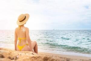 un' giovane caucasico donna nel un' costume da bagno e un' cannuccia cappello si siede su il spiaggia nel davanti di il mare con sua indietro nel il telaio. il sole su un' donna di indietro è disegnato con protezione solare foto