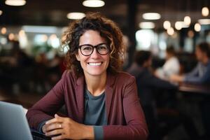 ai generato positivo mezzo anziano donna d'affari utilizzando il computer portatile mentre Lavorando nel coworking spazio, sorridente signora nel bicchieri seduta a tavolo in casa e guardare a telecamera foto