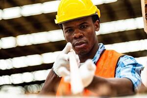 africano lavoratore falegname indossare sicurezza uniforme e difficile cappello Lavorando e controllo il qualità di di legno prodotti a laboratorio produzione. uomo e donna lavoratori legna nel buio magazzino industria. foto