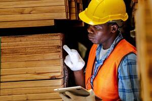 africano lavoratore falegname indossare sicurezza uniforme e difficile cappello Lavorando e controllo il qualità di di legno prodotti a laboratorio produzione. uomo e donna lavoratori legna nel buio magazzino industria. foto