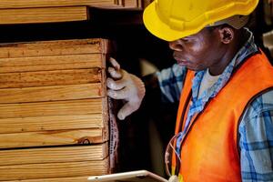 africano lavoratore falegname indossare sicurezza uniforme e difficile cappello Lavorando e controllo il qualità di di legno prodotti a laboratorio produzione. uomo e donna lavoratori legna nel buio magazzino industria. foto