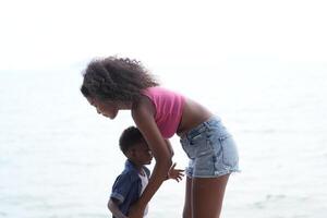 misto gara africano e asiatico madre e ragazzo è giocando a il all'aperto la zona. sorridente contento famiglia avere divertimento in esecuzione su il spiaggia. ritratto di mamma e ragazzo stile di vita con un' unico acconciatura. foto