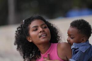 misto gara africano e asiatico madre e ragazzo è giocando a il all'aperto la zona. sorridente contento famiglia avere divertimento in esecuzione su il spiaggia. ritratto di mamma e ragazzo stile di vita con un' unico acconciatura. foto