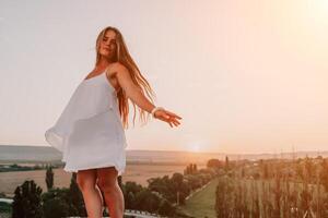 contento donna in piedi con sua indietro su il tramonto nel natura nel estate con Aperto mani. romantico bellissimo sposa nel bianca boho vestito in posa con montagne su tramonto foto