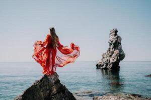 donna viaggio mare. giovane contento donna nel un' lungo rosso vestito in posa su un' spiaggia vicino il mare su sfondo di vulcanico rocce, piace nel Islanda, condivisione viaggio avventura viaggio foto