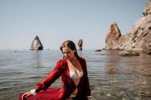 donna viaggio mare. contento turista nel rosso vestito godere assunzione immagine all'aperto per ricordi. donna viaggiatore in posa nel mare spiaggia, circondato di vulcanico montagne, condivisione viaggio avventura viaggio foto