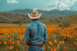 ai generato indietro Visualizza di un' aspro cowboy nel un' denim giacca e cowboy cappello, guardando al di sopra di un' campo di arancia fiori selvatici con montagne nel il distanza foto
