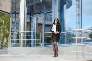 giovane ragazza ufficio lavoratore nel occhiali, buio giacca e blu camicia soggiorni su città strada vicino ufficio edificio e detiene sua documenti. foto