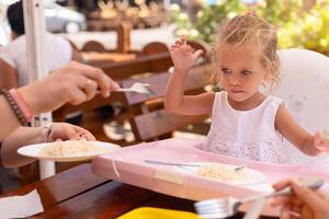 carino poco caucasico ragazza mangiare spaghetti a tavolo seduta nel bambino posto a sedere all'aperto ristorante. foto