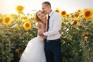 sposa nel nozze bianca vestiti e sposo nel di Pentecoste camicia e cravatta in piedi abbracciare nel il campo di girasoli foto