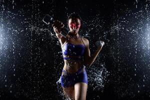 bellissimo giovane ragazza nel viola abbigliamento sportivo pose con manubri nel acqua studio. gocce di acqua diffusione di sua fitness corpo. il Perfetto figura su il sfondo di acqua spruzzi foto