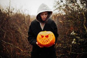 contento Halloween ritratto di nero moderno stile di vita strega nel cappuccio e nero cappotto mantello su di legno cespuglio foresta sfondo. Tenere zucca nel mani foto