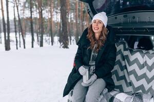 giovane donna nel di lana cappello si siede nel il tronco di il auto e detiene un' tazza di caldo tè nel sua mani foto