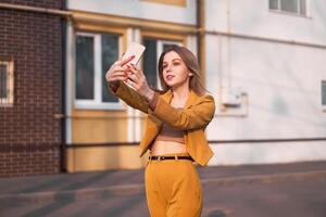 bellissimo giovane caucasico ragazza 20 anni vecchio con bianca capelli vestito elegante arancia attività commerciale completo da uomo fa autoscatto su il Telefono mentre in piedi su il strada. foto