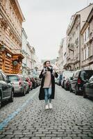 elegante donna nel nero cappotto e sciarpa passeggiando giù ciottolo strada. urbano moda scena con nuvoloso cielo e parcheggiata macchine. città stile di vita concetto con elegante femmina modello all'aperto. foto