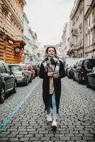 elegante donna nel nero cappotto e sciarpa a piedi su ciottolo strada. urbano moda, città stile di vita concetto - pedone nel paesaggio urbano con parcheggiata macchine, inverno scena, urbano esplorazione e alla moda foto