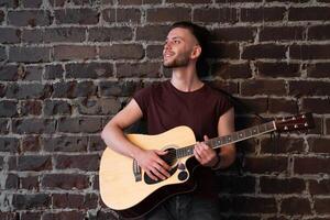 uomo con acustico chitarra contro mattone parete giocando musica cantando canzoni godere vita medio sparare foto