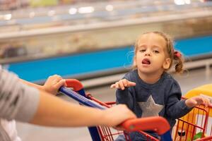 poco ragazza si siede shopping carrello vicino negozi. foto