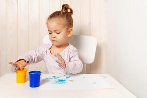 bellissimo poco ragazza disegna con dito vernici su un' bianca foglio di carta. creativo bambino sviluppo nel scuola materna o gratuito tempo a casa foto