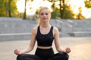 yoga e meditazione nel moderno città caucasico donna rilassare loto posizione seduta yoga stuoia all'aperto estate parco su calcestruzzo pavimento foto