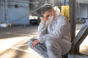 disperazione assistenza sanitaria lavoratore nel bianca covid protettivo tuta da lavoro. stanco maschio caucasico medico afferra il suo testa rannicchiarsi nel depressione durante coronavirus pandemia foto