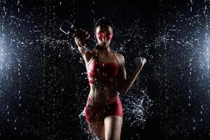 bellissimo giovane ragazza nel rosso abbigliamento sportivo pose con manubri nel acqua studio. gocce di acqua diffusione di sua fitness corpo. il Perfetto figura su il sfondo di acqua spruzzi foto