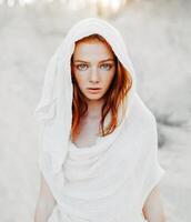 un' bellissimo giovane ragazza con rosso capelli e lentiggini sembra intensamente a il telecamera. donna nel il cappuccio e Abiti per il deserto. concetto foto