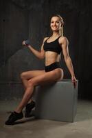 Immagine di muscolare giovane femmina atleta indossare nero sport indossare seduta guardare nel telecamera su buio sfondo. foto