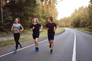 un' gruppo di tre persone atleti uno ragazza e Due uomini correre su un asfalto strada nel un' pino foresta. foto