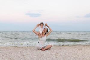 contento famiglia madre e bambino figlia fare yoga, meditare nel loto posizione su spiaggia a tramonto foto