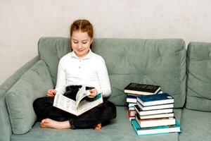 poco ragazza lettura un' libro seduta su il divano foto