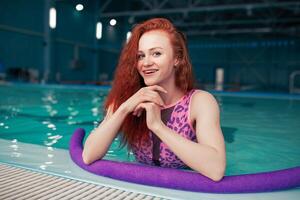 bellissimo giovane dai capelli rossi ragazza nel un' alla moda costume da bagno nel il stile di il anni 80 sta con il tagliatelle nel sua mani nel il interno nuoto piscina foto