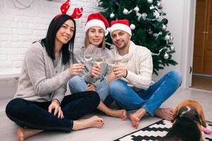 contento amici festeggiare nuovo anno nel casa interno nel Natale cappelli seduta vicino un' Natale albero con bicchieri di vino foto