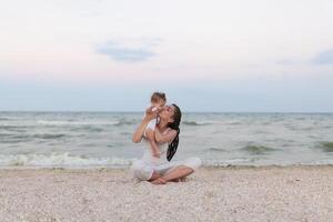 contento famiglia madre e bambino figlia fare yoga, meditare nel loto posizione su spiaggia a tramonto foto