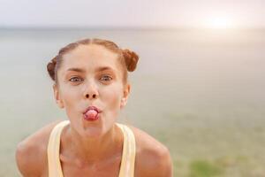 ritratto di giovane elegante ridendo ragazza modello estate naturale trucco al di fuori su il spiaggia. guardare a telecamera mostrando sua lingua foto