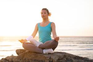 donna pratica yoga loto posa per meditazione con estate vacanza roccioso spiaggia felicità e rilassamento. calma femmina esercizio con yoga meditare oceano spiaggia con tramonto d'oro volta. foto