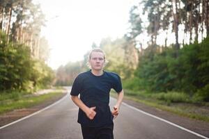 caucasico giovane Riccio uomo atleta corre soleggiato estate giorno su asfalto strada nel il foresta. foto