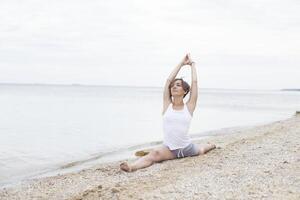 bellissimo ragazza praticante yoga su il spiaggia vicino il mare. si siede su un' spago, fa un' allungamento. foto