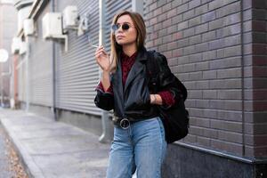 bellissimo giovane con stile vestito caucasico ragazza fuma sigaretta su il strada. foto