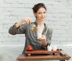 orientale maestro di tè cerimonia con bianca mattone parete su il sfondo. tradizionale tè festa su il natura con donna vestito nel il grigio camicia foto