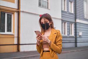 giovane bellissimo caucasico ragazza 20 anni indossare nero viso maschera protezione contro epidemico coronavirus covid-19 passeggiate giù il strada sembra a il Telefono schermo. foto