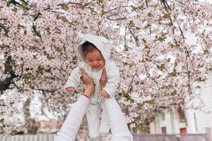 ritratto di contento gioioso bambino nel bianca Abiti al di sopra di albero fiori fiorire sfondo. famiglia giocando insieme fuori. mamma allegramente hold poco figlia foto