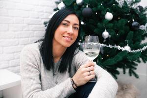 un' giovane donna si siede vicino un' Natale albero, Tenere un' bicchiere e un' Natale giocattolo. preparazione per il nuovo anni celebrazione foto