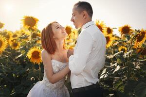 sposa nel nozze bianca vestiti e sposo nel di Pentecoste camicia e cravatta in piedi abbracciare nel il campo di girasoli foto