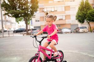 carino poco sorridente ragazza equitazione bicicletta bicicletta nel città su parcheggio soleggiato estate giorno. attivo famiglia tempo libero con bambini. foto