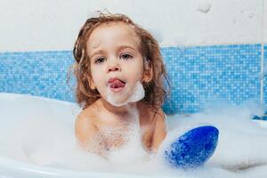 poco bellissimo ragazza bagna nel un' bagno, giochi il scemo e Spettacoli lingua. divertimento bagnarsi bambini. positivo igiene. indipendente bambino foto