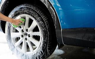 un' persona è lavaggio un' auto pneumatico con un' verde spazzola. pneumatico è coperto nel acqua e sapone. blu auto lavare con bianca sapone schiuma. auto cura servizio. auto pulizia servizio concetto. veicolo pulizia servizio. foto