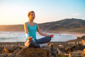 donna pratica yoga loto posa per meditazione con estate vacanza roccioso spiaggia felicità e rilassamento. calma femmina esercizio con yoga meditare oceano spiaggia con tramonto d'oro volta. foto